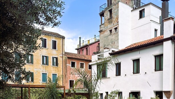 An outdoor seating area with wicker furniture and a wooden table, surrounded by greenery and overlooking a courtyard with buildings. - U-Visionary Venezia Hotel (Venice)