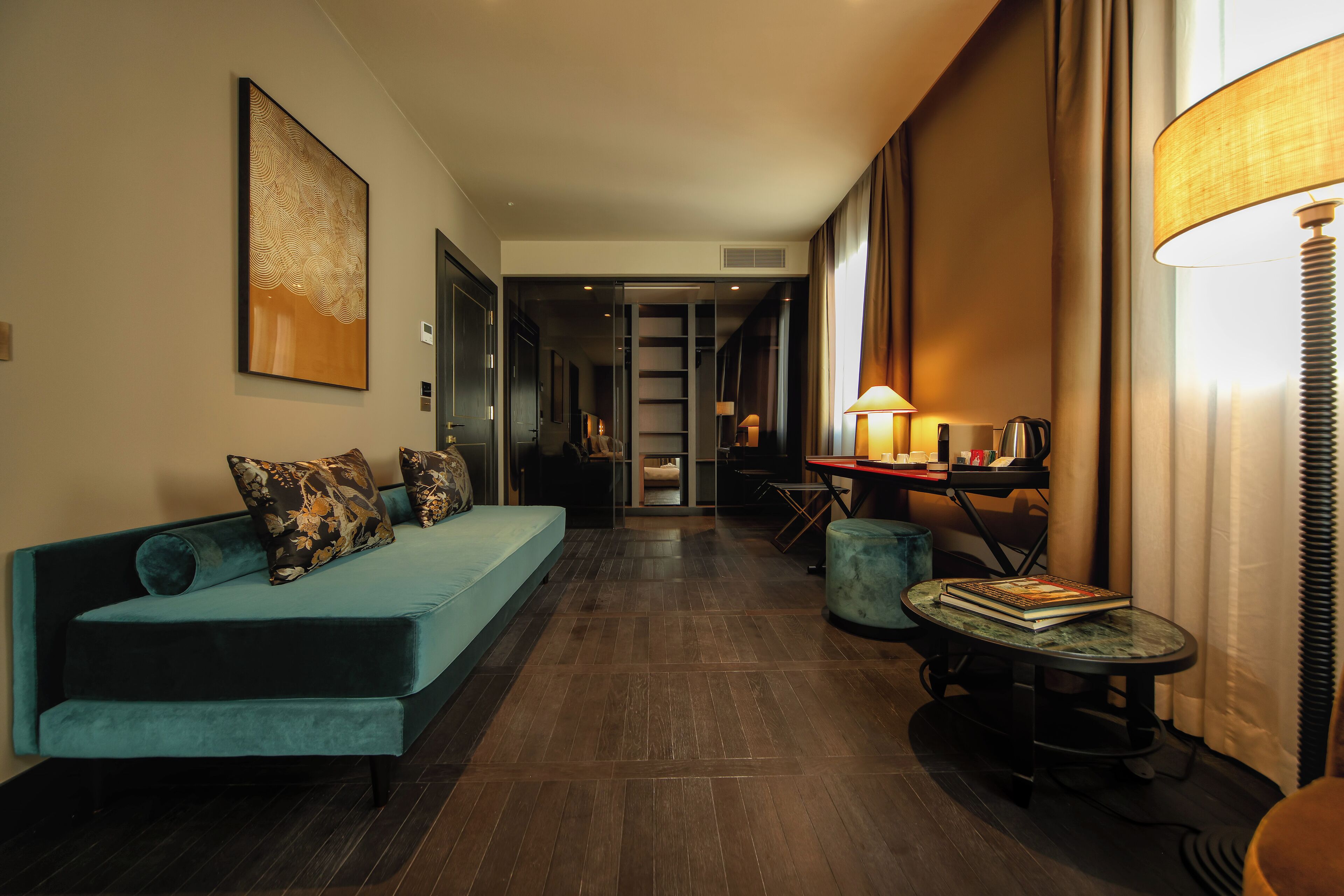 Un moderno hall de hotel con un sofá color turquesa, una pequeña mesa redonda con una lámpara y un cuadro abstracto grande en la pared.
