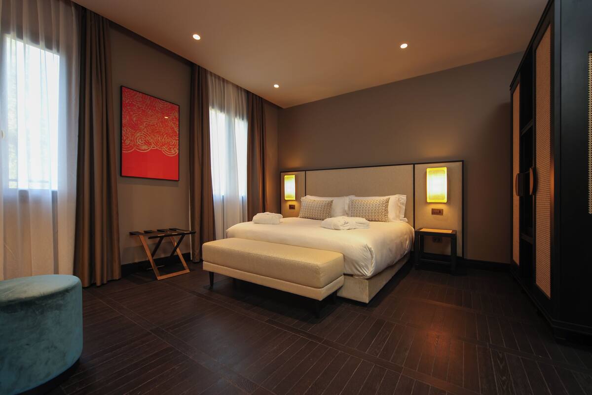 Een moderne hotelkamer met een groot bed, nachtlampjes, een tafeltje en een bankje.