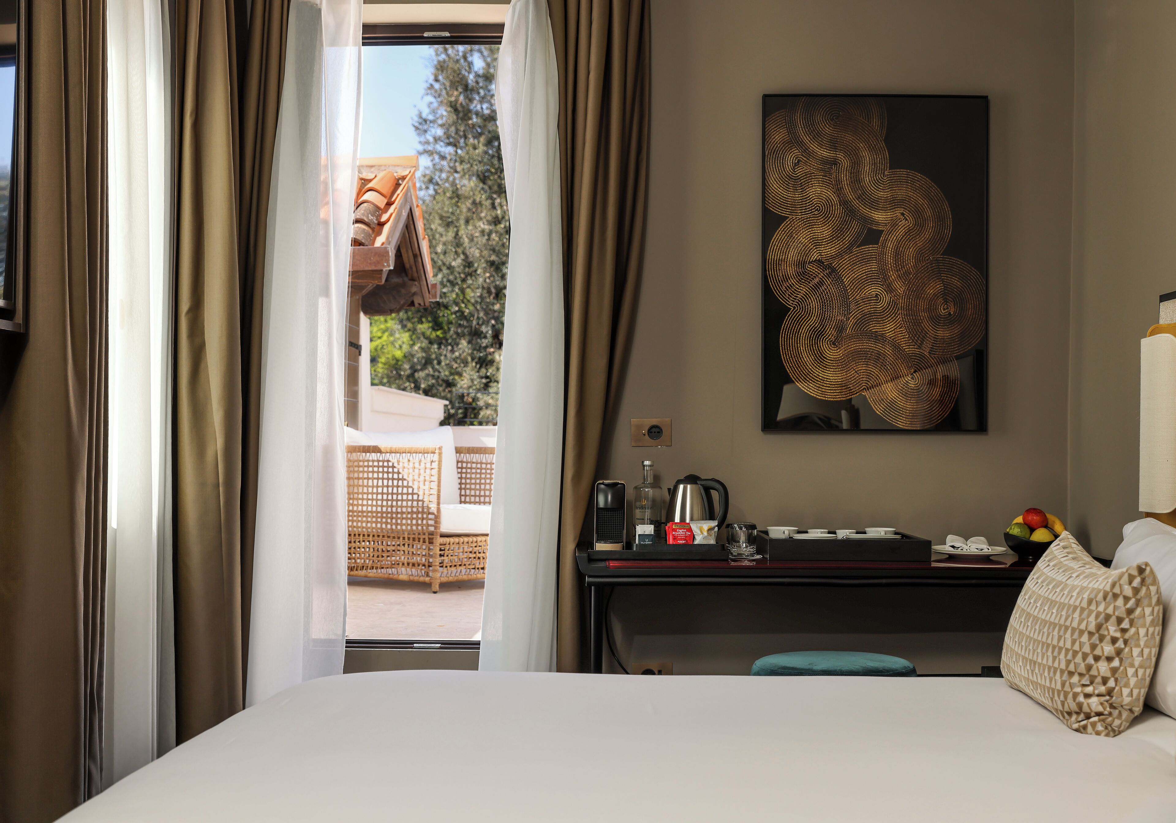 A hotel room with a bed, a desk with a coffee maker, a bowl of fruit, and a painting on the wall.