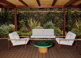 A covered outdoor seating area with a wooden deck, a round wooden table, and a grey sofa with armchairs.