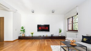A modern living room with a wooden floor, a flat-screen TV mounted on the wall, a sofa, a coffee table, and a window with a view of trees.
