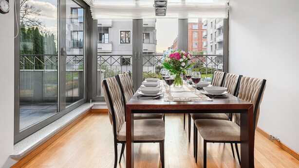 A dining area with a wooden table set for four, surrounded by cushioned chairs, with a view of a balcony and buildings in the background.