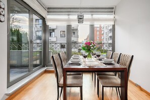 A dining area with a wooden table set for four, surrounded by cushioned chairs, with a view of a balcony and buildings in the background. - Lumina stylish apartment with parking (Lódz)