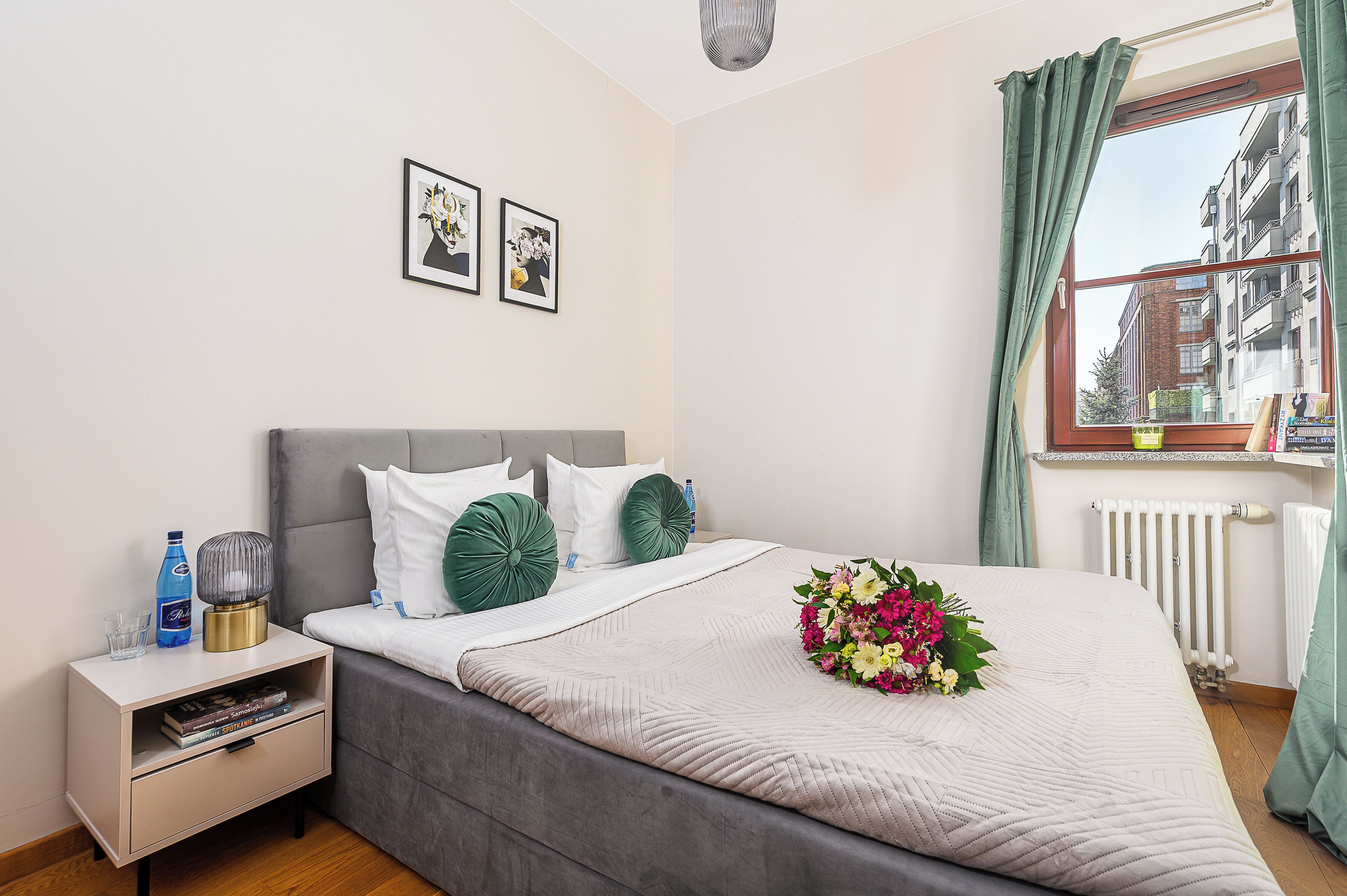 Een keurig opgemaakt slaapkamer met een bed, nachtkastje, raam met gordijnen en twee ingelijste foto's aan de muur.