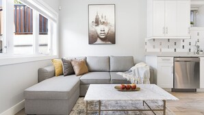 A modern living room with a grey sectional sofa, a white coffee table, and a cityscape artwork on the wall. - Cozy & Convenient · 2 Mins from SkyTrain · Central Location (Vancouver)