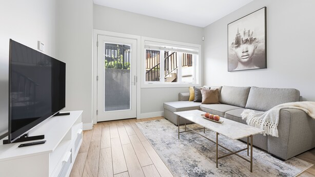 A modern living room with a flat-screen TV, a grey sofa, a coffee table, and a large window with a view of a cityscape. - Cozy & Convenient · 2 Mins from SkyTrain · Central Location (Vancouver)