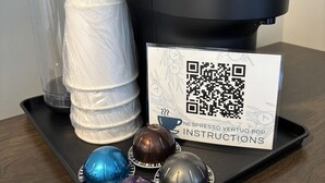Uma máquina de café Nespresso Vertuo Pop, uma pilha de copos de papel brancos, um código QR e quatro cápsulas de café em azul, castanho, cinza e roxo.