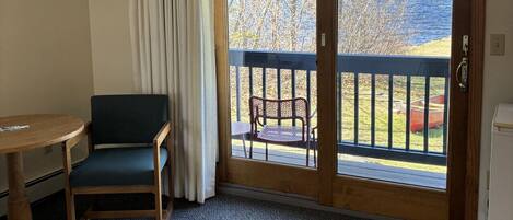 A room with a wooden chair and a small table, a sliding glass door leading to an outdoor deck with a view of the water and trees.