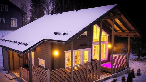 Eine Holzhütte mit schneebedecktem Dach, großen Fenstern und einer Veranda mit Beleuchtung.
