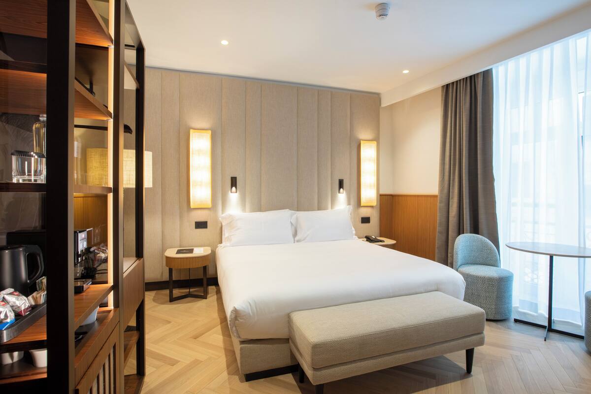Ein modernes Hotelzimmer mit einem großen Bett, Nachttischen, einer Sitzecke und Blick auf die Stadt durch große Fenster.