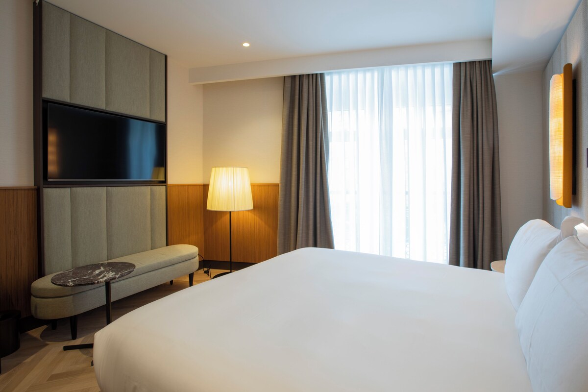 Hotelkamer met een groot bed, een flatscreen televisie aan de muur, een bankje met tafel, een lamp en een raam met gordijnen.