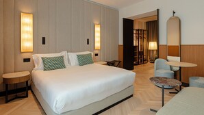 Une chambre d’hôtel moderne dotée d’un grand lit, de deux tables de chevet, d’une table basse ronde et d’un coin salon avec un fauteuil et une lampe.