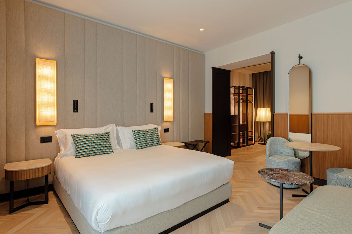 Ein modernes Hotelzimmer mit einem großen Bett, zwei Nachttischen, einem runden Couchtisch und einer Sitzecke mit Sessel und Lampe.