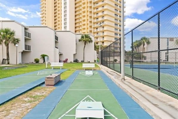 Een tennisbaan met net, omringd door een hek, naast een gebouw met plat dak en meerdere ramen.