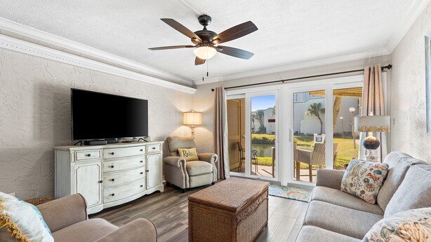 A living room with a flat-screen TV, a ceiling fan, a sofa, a wicker ottoman, and a sliding glass door leading to an outdoor patio. - 1BR Ground Floor, Pets, Beach & Pool, Sleeps 4 (Panama City Beach)