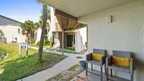 A covered outdoor seating area with wicker chairs and a yellow cushion, a sign prohibiting fishing, and a building with a balcony in the background. - 1BR Ground Floor, Pets, Beach & Pool, Sleeps 4 (Panama City Beach)