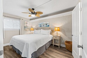 A bedroom with a bed, two bedside tables, a ceiling fan, a window with blinds, and a wall with a painting.