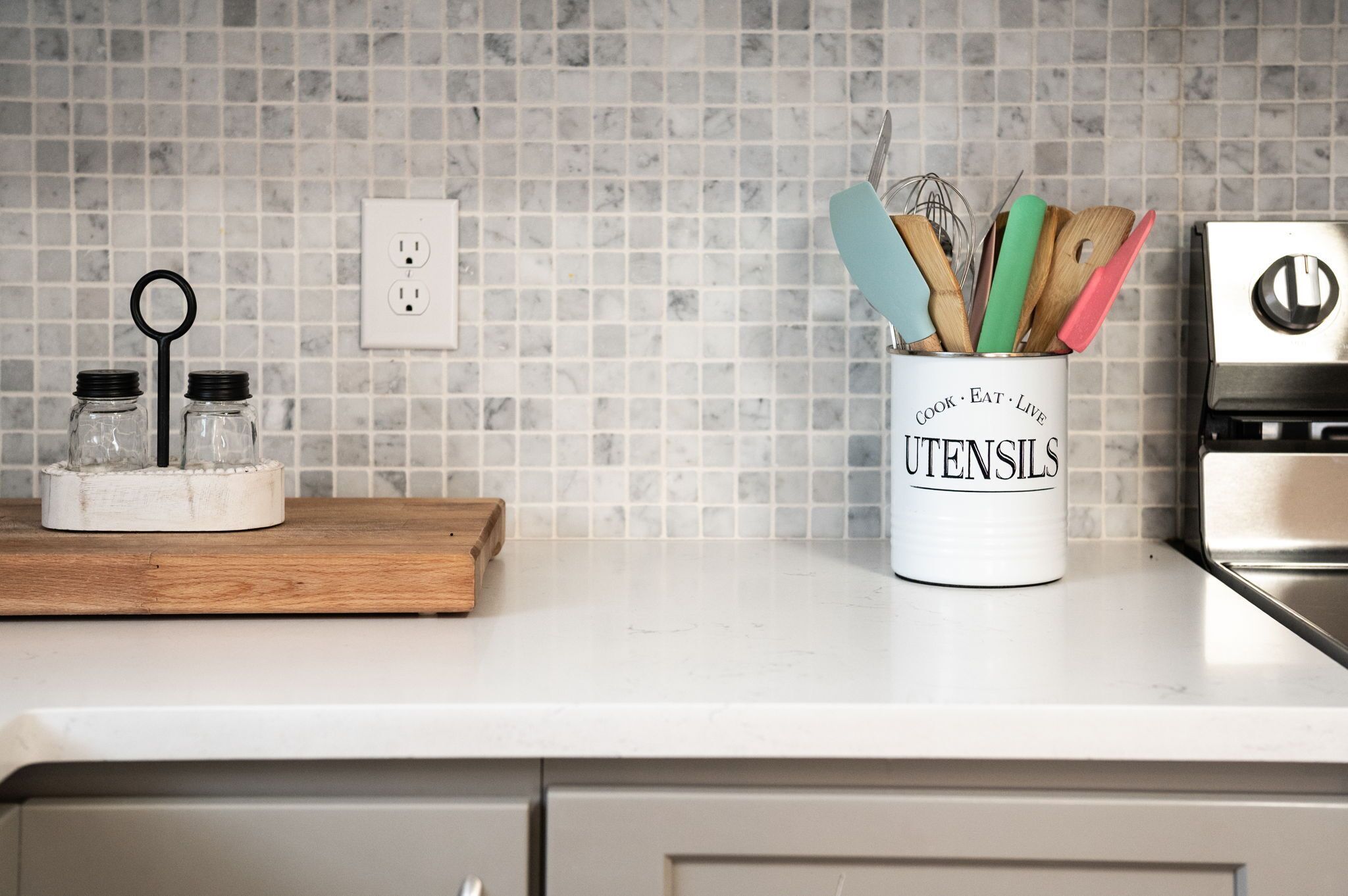 Una encimera de cocina con un utensilero blanco etiquetado “Cook Eat Live Utensils” y una tabla de cortar de madera.
