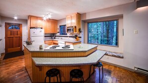 Une cuisine avec des meubles en bois, un îlot central doté de tabourets, un réfrigérateur et une grande fenêtre donnant sur une forêt.