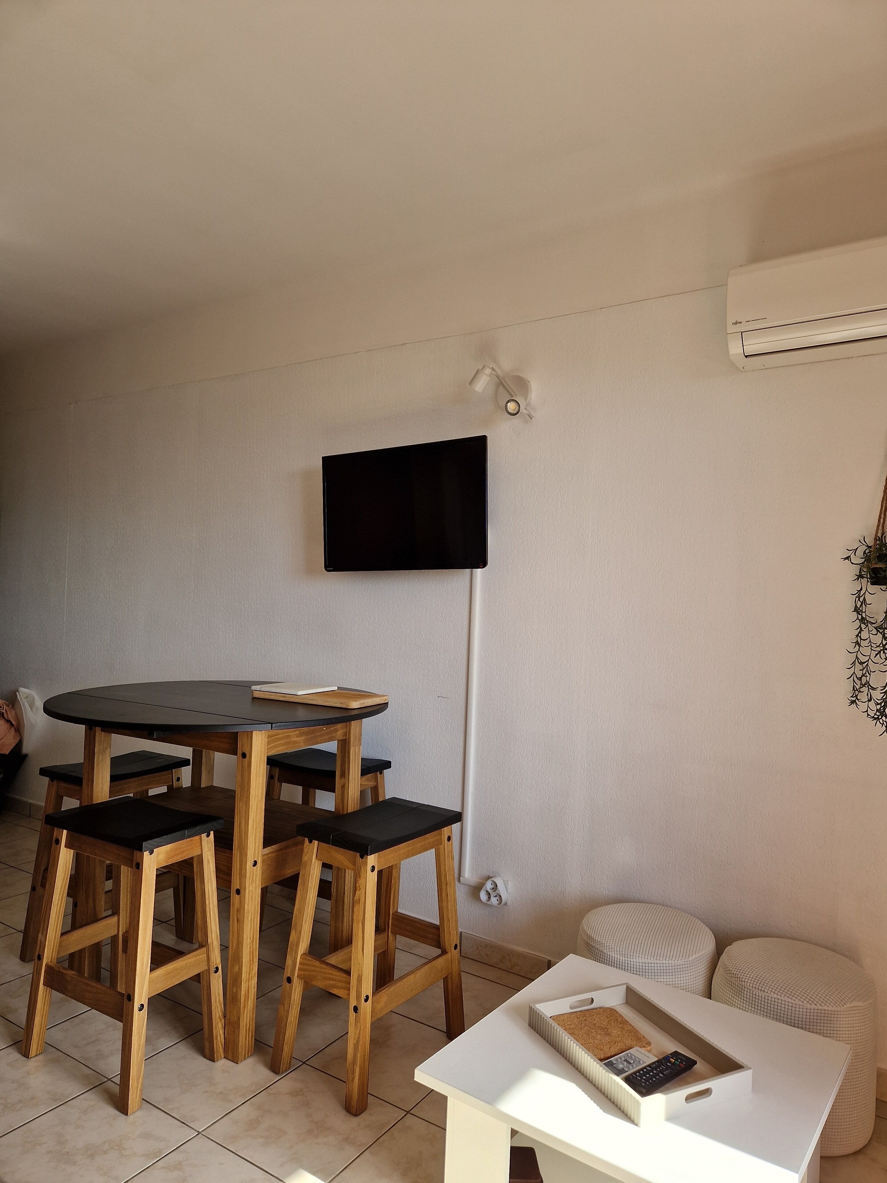 Un coin repas avec une table ronde et des chaises en bois, un téléviseur à écran plat fixé au mur et une petite table d’appoint blanche avec une télécommande.