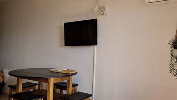 Un coin repas avec une table ronde et des chaises en bois, un téléviseur à écran plat fixé au mur et une petite table d’appoint blanche avec une télécommande.