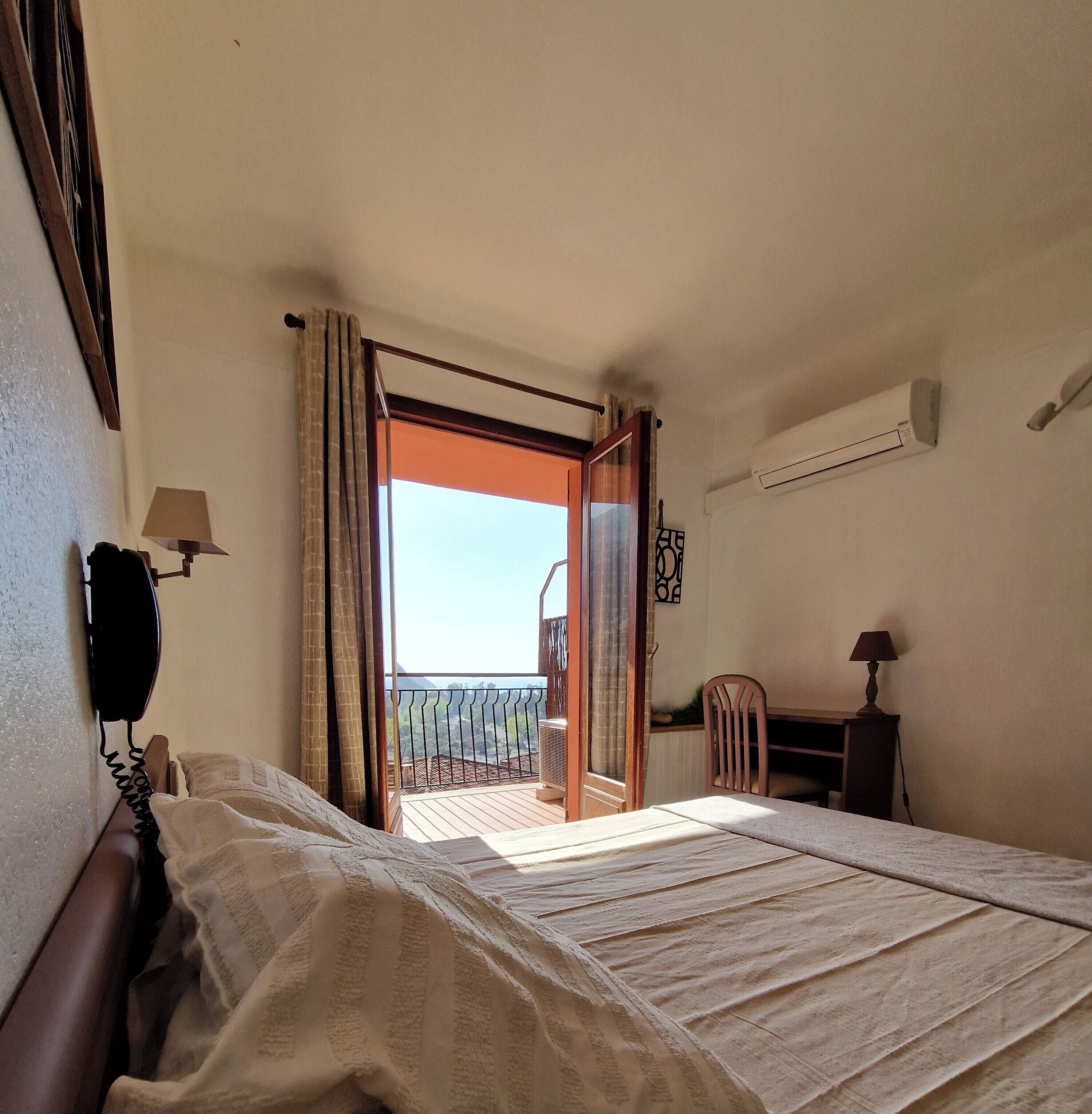 A hotel room with a bed, a desk, a chair, a telephone, and an air conditioning unit.