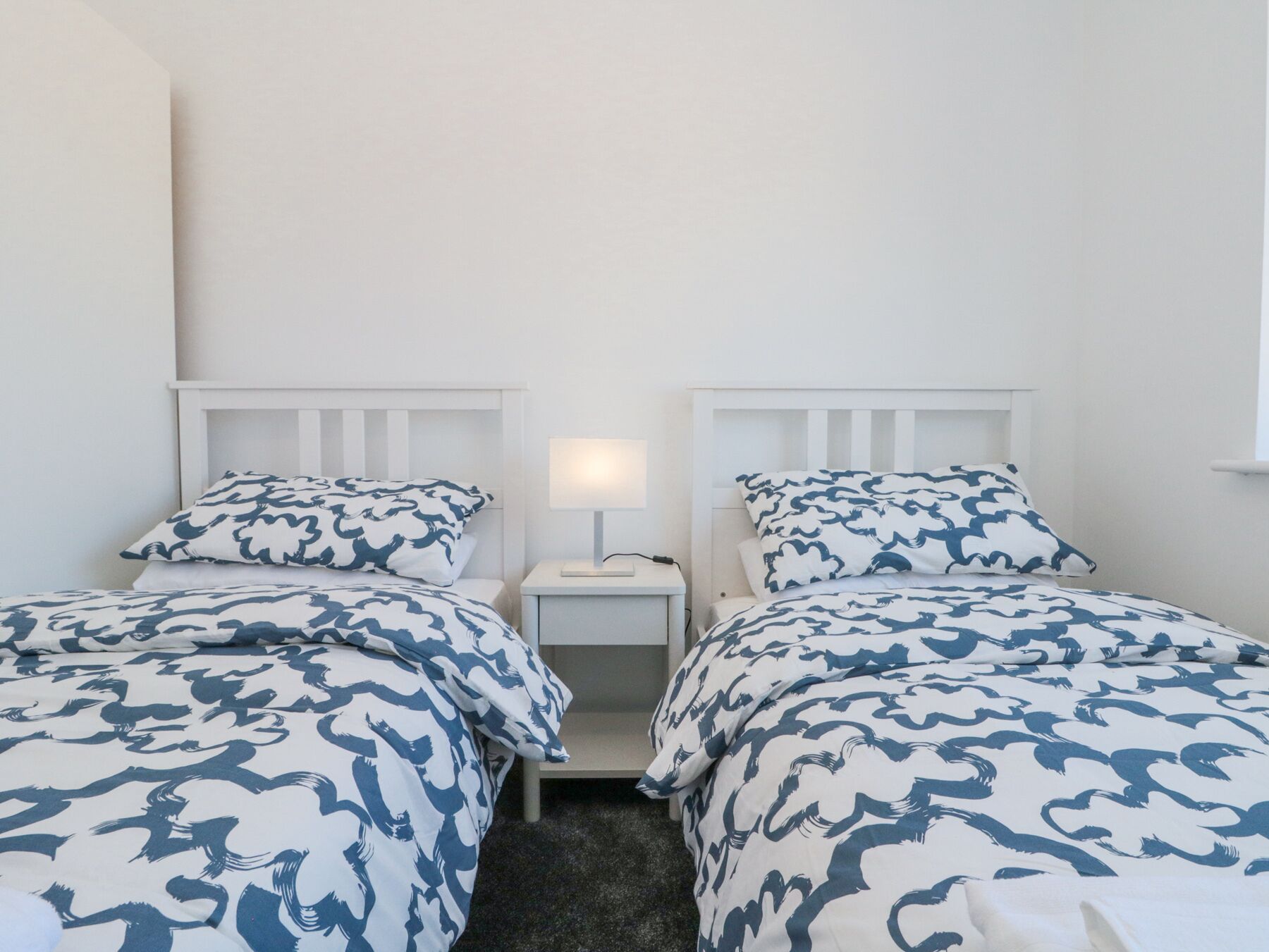 A room with two single beds, each with a white headboard and matching pillowcases. There is a bedside table with a lamp between the beds.