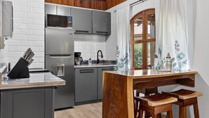 A compact kitchen with grey cabinets, a stainless steel refrigerator, a microwave, and a coffee maker. There's a wooden dining table with two stools. - Tropical Perfection, Includes Kayak, Snorkel, Beach Gear, Walk To The Beach (Playa Potrero)