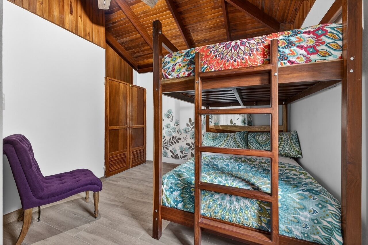 Ein Stockbett mit bunter Bettwäsche, ein Holzstuhl und ein Kleiderschrank in einem Raum mit Holzbalken und Fliesenboden.