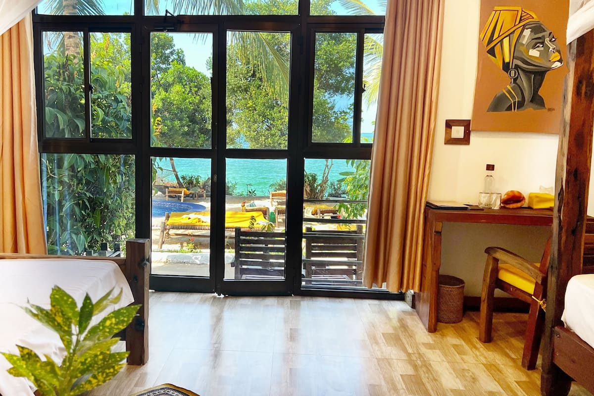 A room with a large window overlooking a beach, a bed, a desk, and a chair.