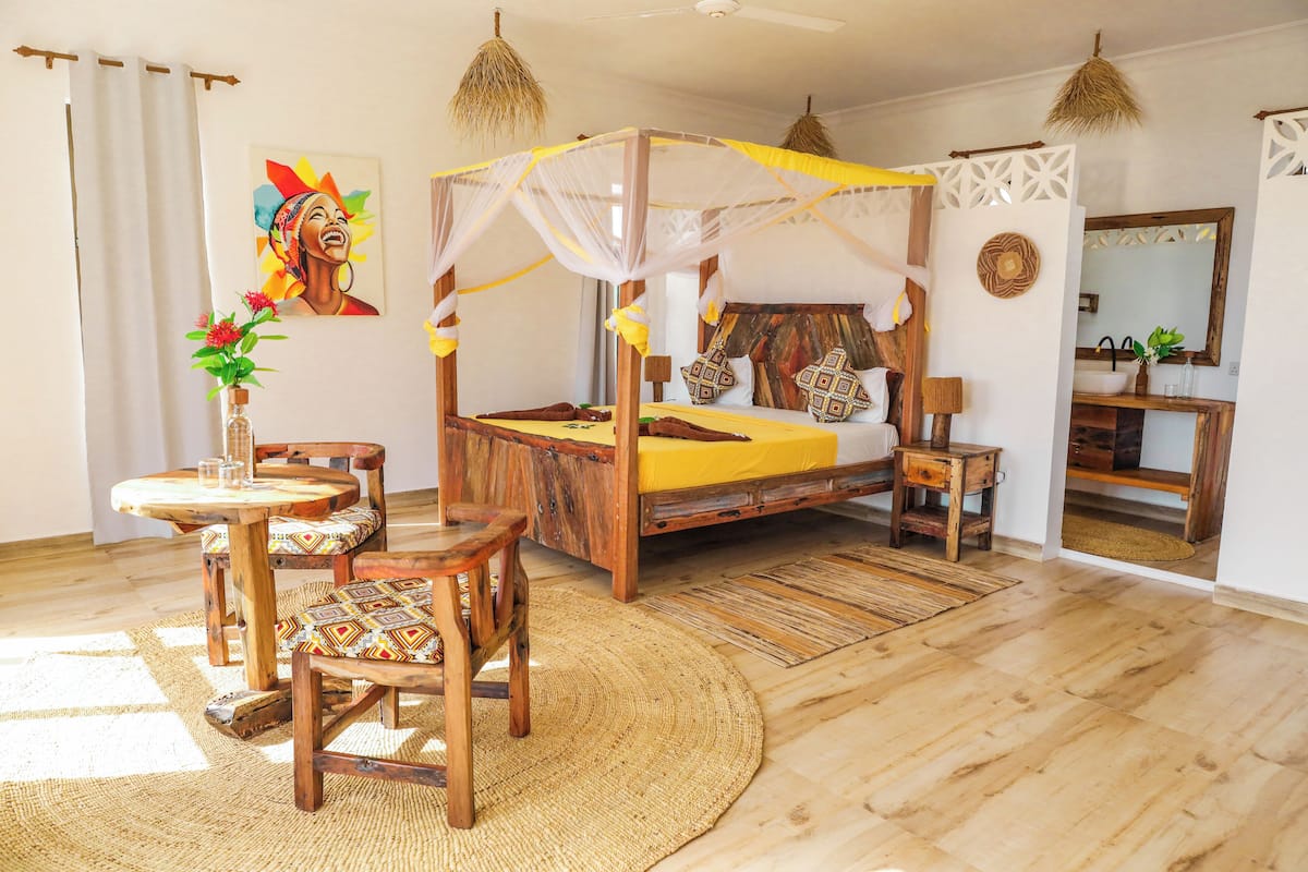 A room with a wooden four-poster bed, a round wooden table with chairs, a woven rug, and a painting on the wall.