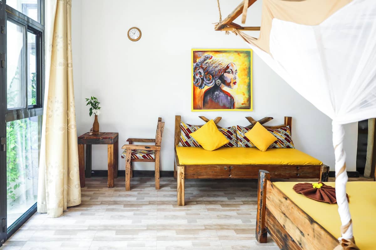 A room with a wooden bed, yellow pillows, a small wooden table, a chair, a potted plant, and a painting on the wall.