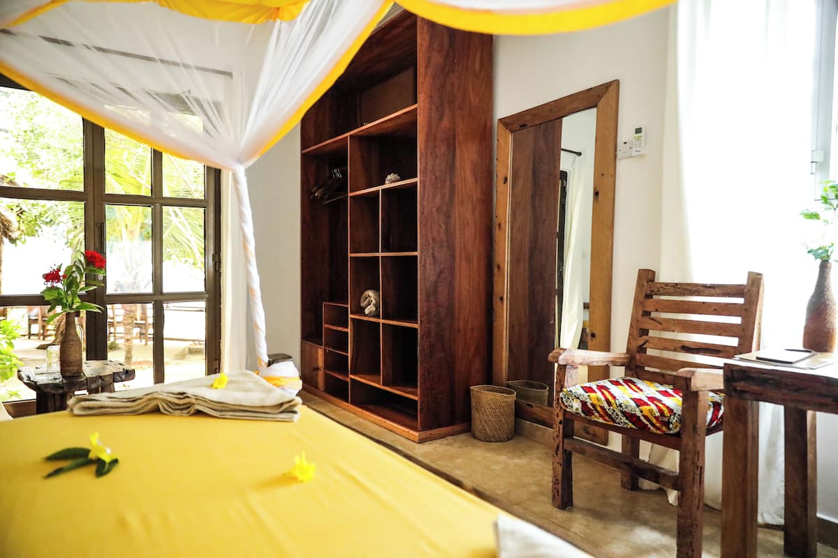 A room with a bed, a wooden chair, a small table, and a window with a view of greenery.