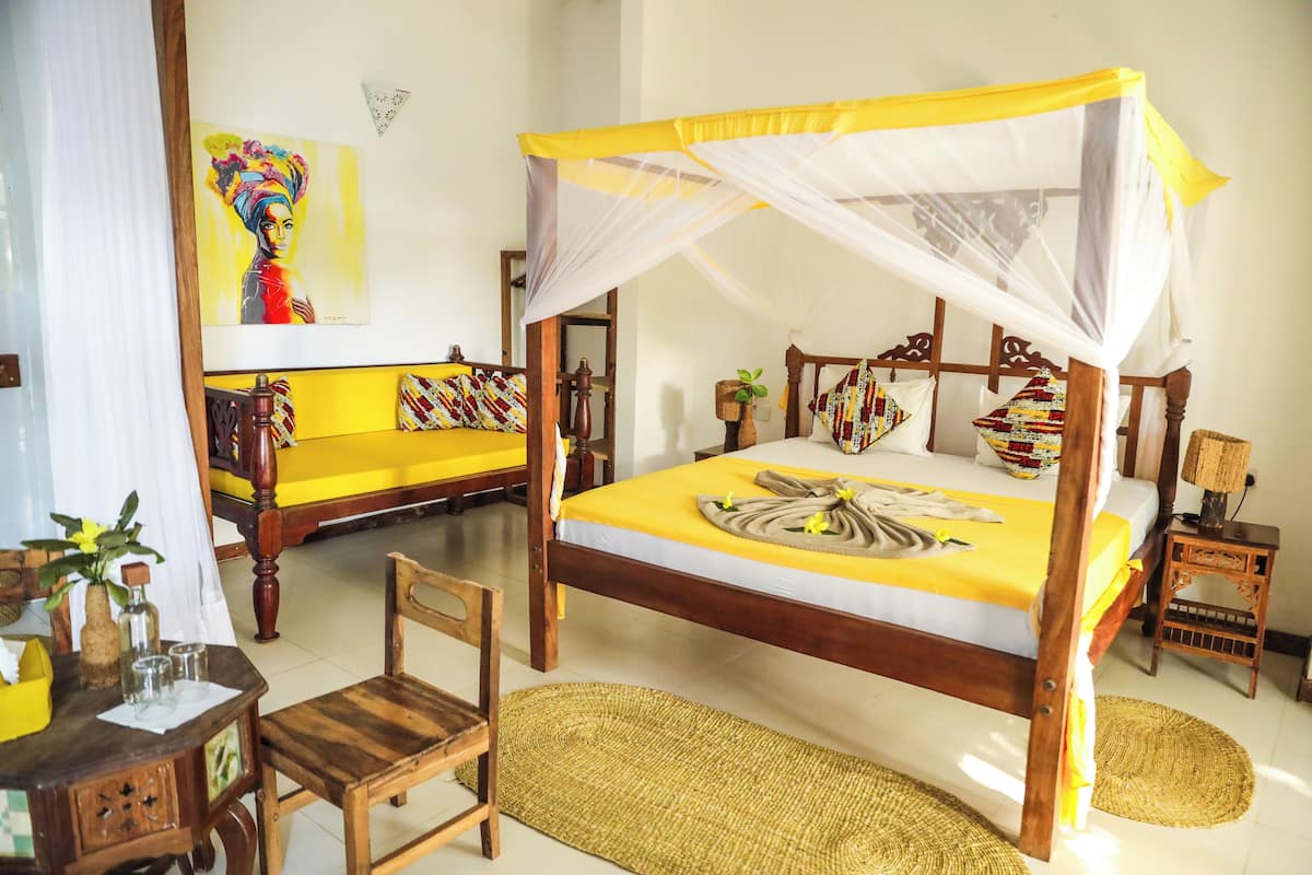 A room with a four-poster bed, a wooden chair, a small table with a vase, and a painting on the wall.