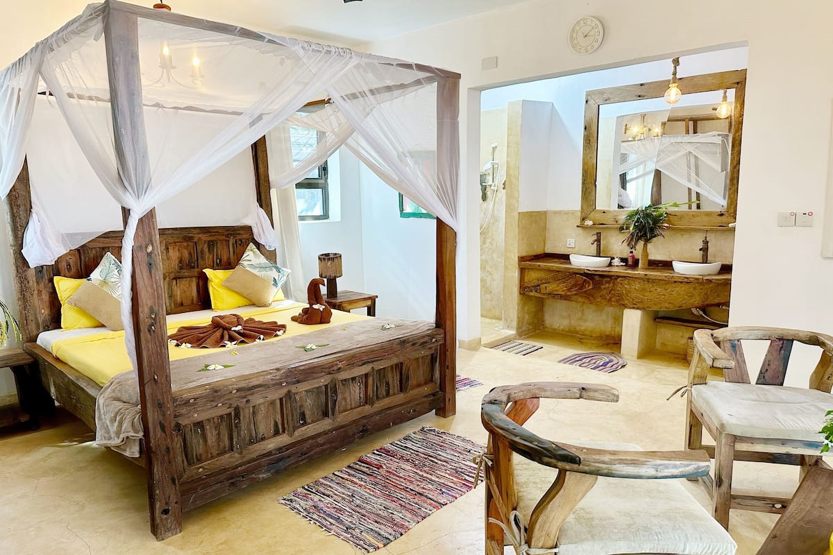 A rustic wooden bed with a canopy, a wooden chair, a small table, and a bathroom with a wooden vanity and mirror.