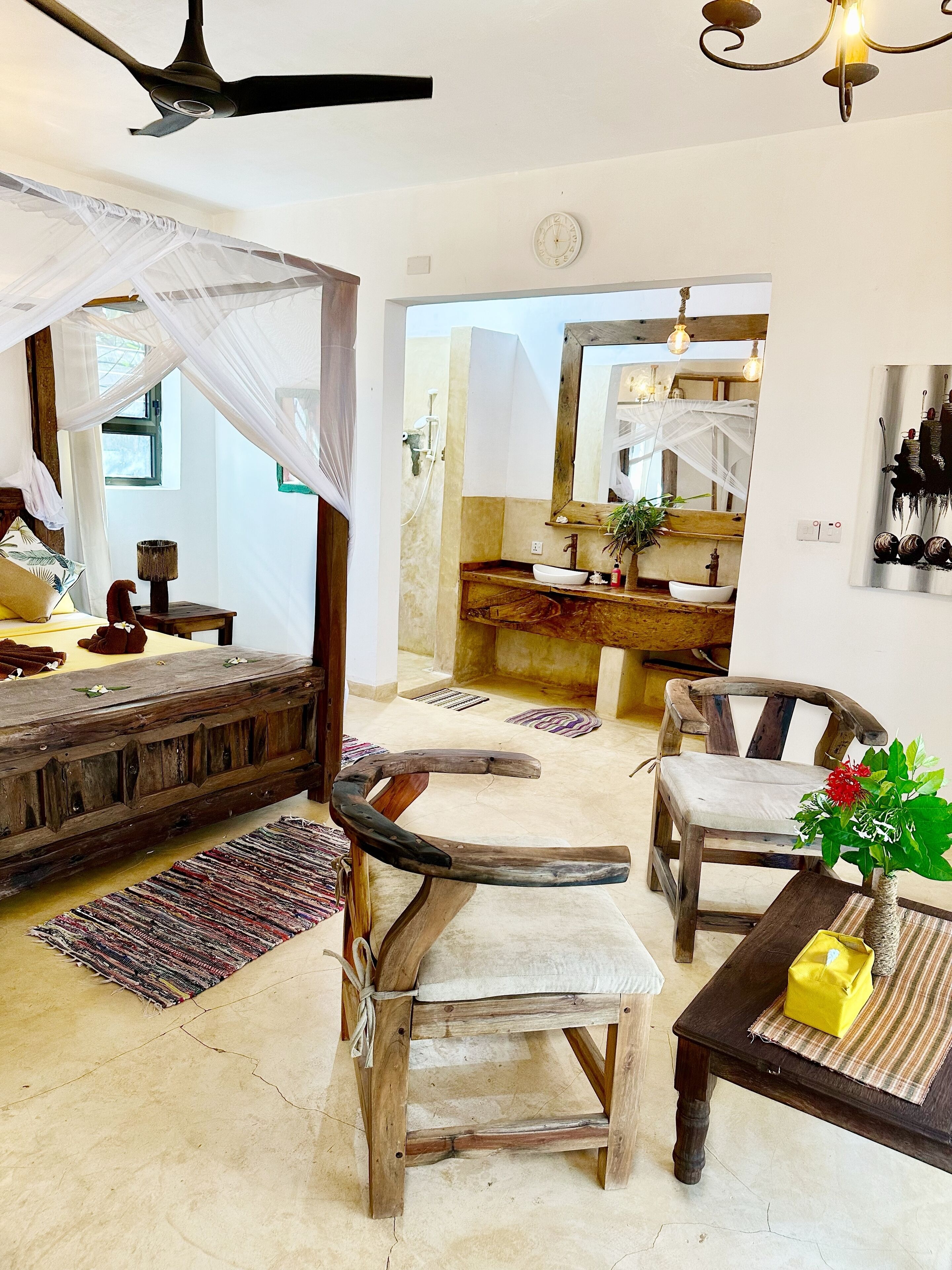 Una habitación con cama, una silla de madera, una mesa, ventilador de techo y un baño visible a través de una puerta abierta.