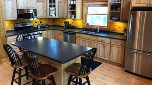 A modern kitchen with wooden cabinets, a stainless steel refrigerator, and a central island with chairs. - 15min to Pictured Rocks|Secluded Cabin w/Sauna (Wetmore)