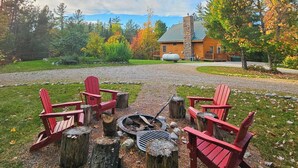 A cozy outdoor seating area with red Adirondack chairs around a fire pit, set against a backdrop of a stone building and autumn foliage. - 15min to Pictured Rocks|Secluded Cabin w/Sauna (Wetmore)