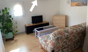 A small, well-lit room with a wooden floor, a sofa, a TV on a wooden stand, a small table, a dresser, and a potted plant.