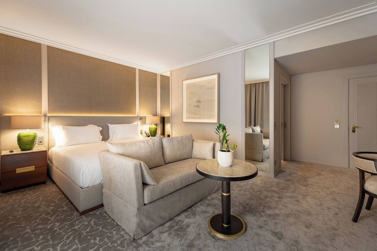 A modern hotel room with a bed, a sofa, a small round table with a plant, a nightstand, and a mirror.