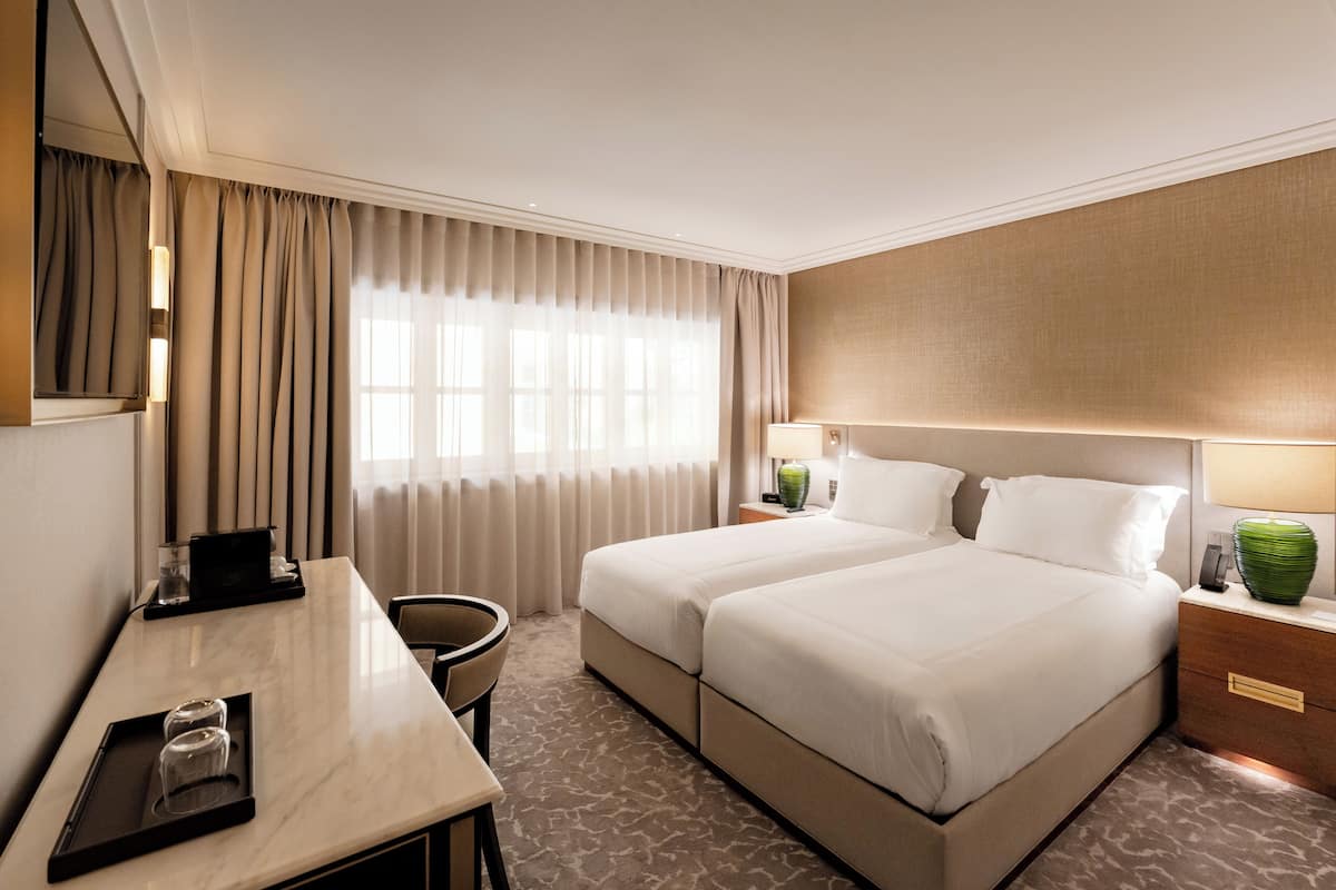 A hotel room with a bed, a desk, a chair, a lamp, and a vase.