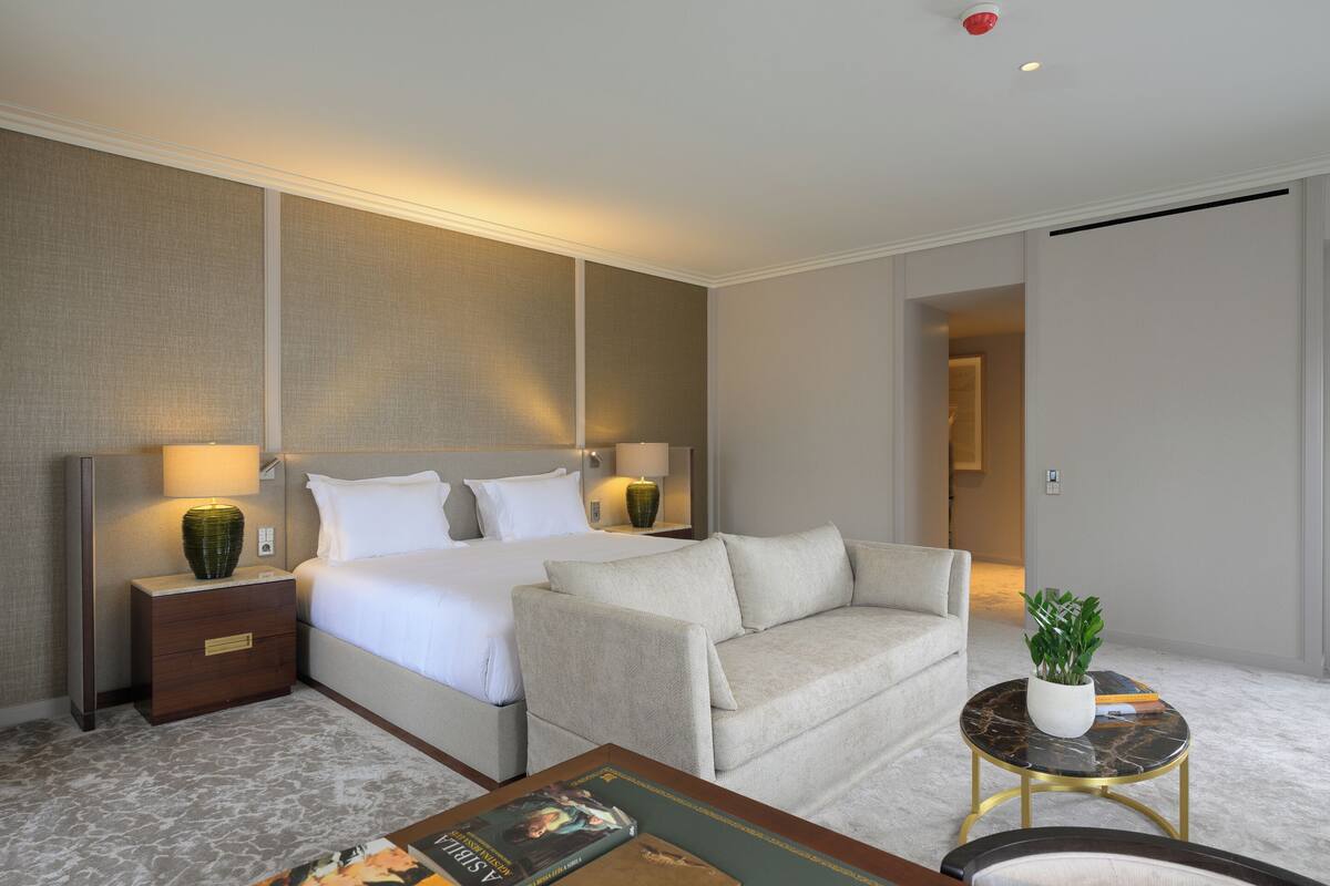 A hotel room with a bed, a sofa, a small round table with a plant, and a nightstand with a lamp.