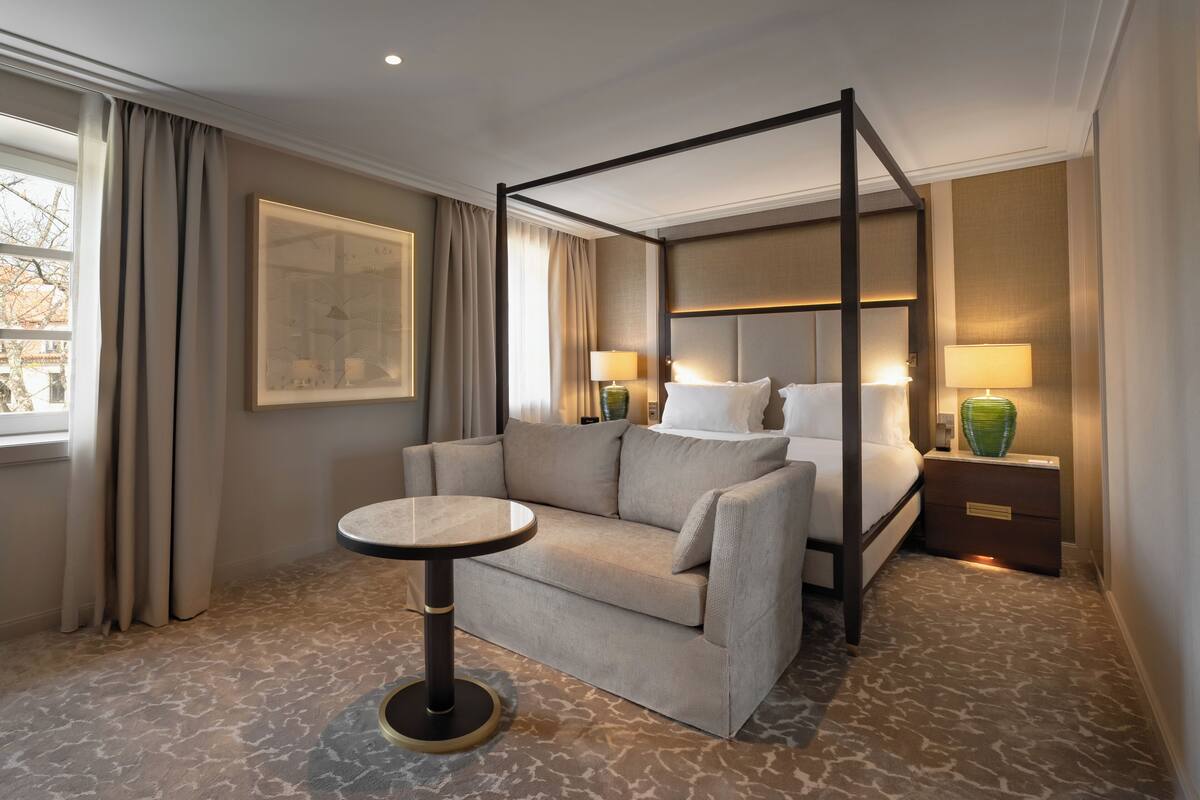 A four-poster bed with a headboard, a sofa, a round table, and a nightstand with lamps.