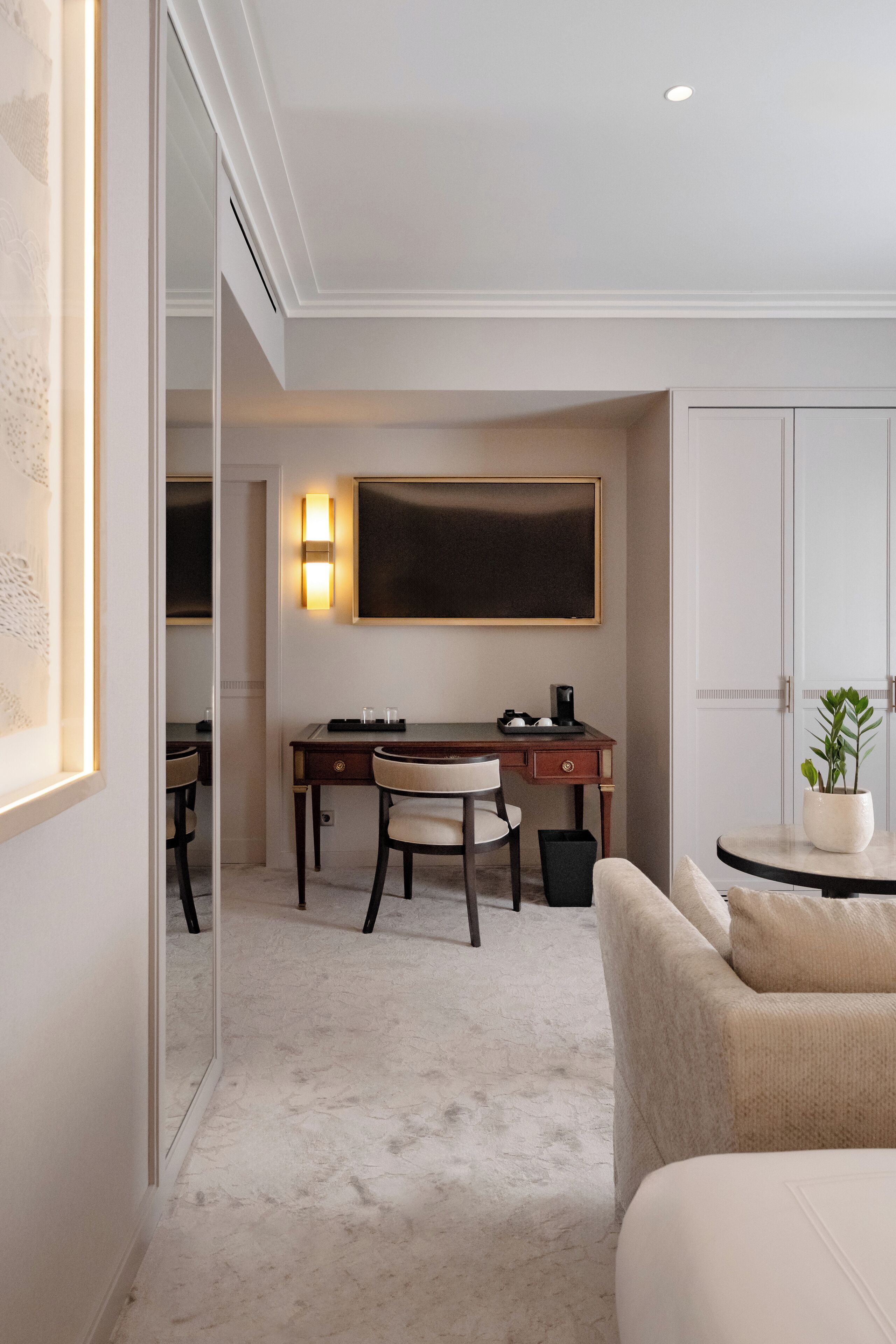 A hotel room with a bed, a desk with a chair, a wall-mounted light, and a potted plant.