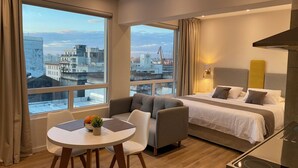 Habitación de hotel moderna con cama, sofá, mesa de comedor y ventanas amplias con vista a la ciudad.