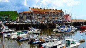 Un port où sont amarrés divers bateaux, un bâtiment avec un toit rouge et, au loin, une colline verdoyante.