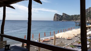 A beach view with umbrellas and tables under a wooden structure, overlooking the sea and a rocky coastline. - Hotiday Room Collection - Nerano (Marina del Cantone)