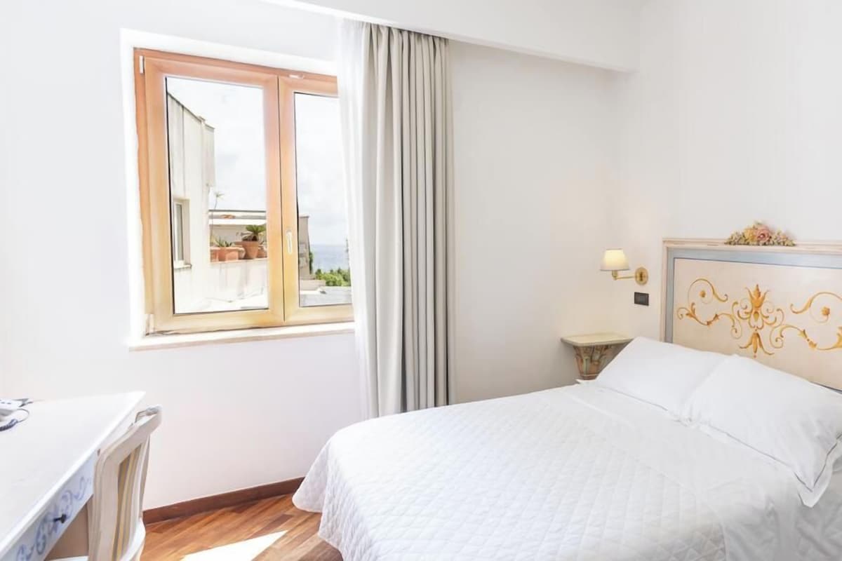 A hotel room with a bed, a desk, a chair, a window with curtains, and a view of the outside.