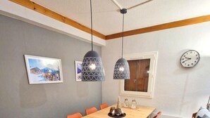 A dining area with a wooden table, orange chairs, pendant lights, a framed picture, and a clock on the wall.